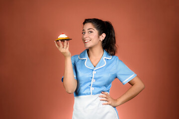 Waitress dressed in a blue uniform. Looking sideways smiling at the camera, offering a cake with one hand and the other on her hip.
