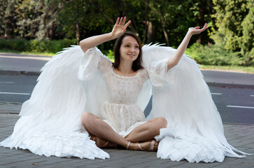 A girl in a white dress and with large white wings sits on the asphalt