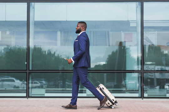 Young african businessman with suitcase and smartphone in hands walking in airport terminal, handsome african american male going to flight boarding, ready for air trip, side view with copy space
