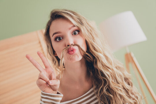Playful young woman with a faux mustache poses with peace sign in a bright living room during the day - Powered by Adobe