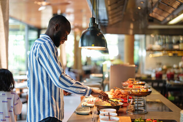 A Black man in a striped shirt picks up food at a self-service buffet in a modern restaurant. The...