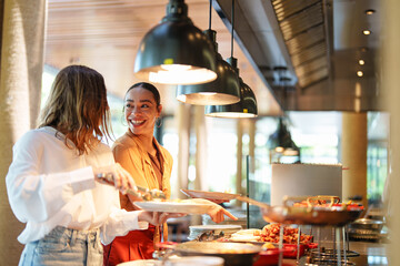 Multiracial and Caucasian young adult females picking up food in a self-service restaurant. One...