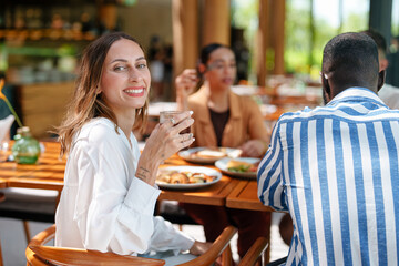 A joyful young Caucasian woman enjoys breakfast with diverse friends at a cozy cafe. The group shares a meal at a wooden table, creating a warm, friendly atmosphere in the brightly lit space.
