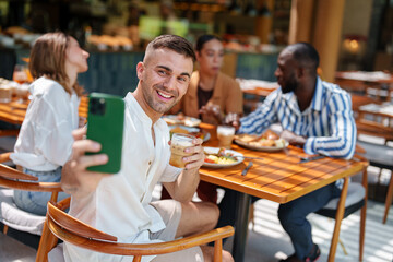 A young Caucasian male joyfully takes a selfie while enjoying breakfast with diverse friends at a cafe. The social gathering features mixed ethnicities, capturing camaraderie.