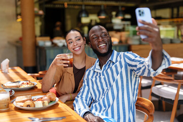 A diverse group enjoys breakfast at a cozy cafe. A young Black male and multiracial female couple take a cheerful selfie, surrounded by friends, warm lighting, and delicious food.