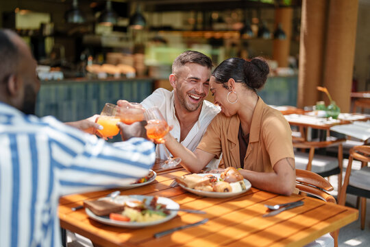 Loving Caucasian male and multiracial female couple share a joyful toast with diverse friends, enjoying fresh juice during a breakfast gathering in a cozy bar.