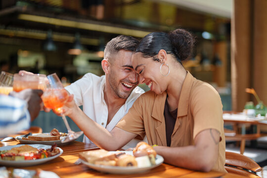 A joyful young adult Caucasian male and a diverse female share a breakfast with friends at a cafe. They are laughing and toasting over a table filled with breakfast dishes, creating a warm atmosphere.