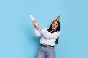 Happy woman in conical paper hat blowing up confetti popper on light blue background. Surprise party