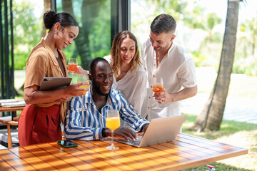 A group of diverse young adult entrepreneurs collaborate on a project at a sunny tropical bar. Engaged in discussion, they use a laptop and digital tablet, surrounded by palm trees and cocktails.