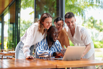 A group of diverse young adult friends joyfully gathers around a laptop at a tropical bar....