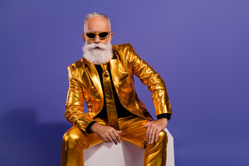 Photo portrait of fashionable old showman sit white cube wear glamour golden garment isolated on violet color background
