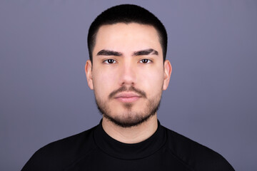 Neutral expression headshot of a young man with short black hair, wearing a black high-collar shirt, posing against a gray background.