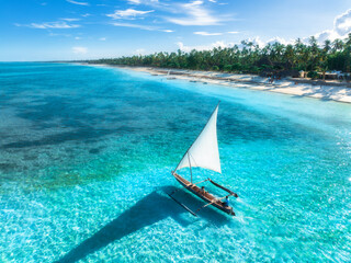 Obraz premium Aerial view of the sailboat on blue sea, empty white sandy beach at sunrise. Summer vacation in Zanzibar. Tropical landscape with boat, ocean with clear water, green palms, sky. Top drone view. Exotic