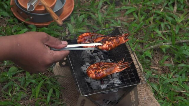 Close up of the activity of an Asian male adventurer cooking grilled shrimp using charcoal and wood. The bushcraft concept makes grilled shrimp with simple equipment in the lush wilderness