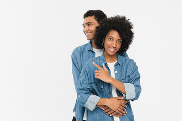 Smiling young african american couple two friends man woman in denim casual clothes posing hugging pointing finger at copy space looking camera isolated on white color wall background studio portrait