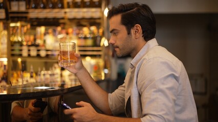 Smart man drinking cocktail in Old fashioned waiting friends or couple while chatting on smartphone to wait at counter bar with bartender preparing drinks at nightclub Friday meeting night. Vinosity.