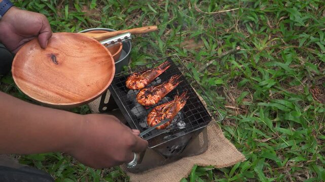 Close up of the activity of an Asian male adventurer cooking grilled shrimp using charcoal and wood. The bushcraft concept makes grilled shrimp with simple equipment in the lush wilderness
