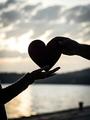 A silhouette of two hands exchanging a heart-shaped object, set against a tranquil lakeside backdrop during sunset.