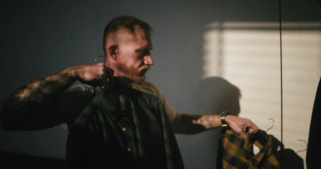 A blond man with a beard in a white T-shirt brings two shirts to himself and tries on choosing one of them Before going to work in the morning