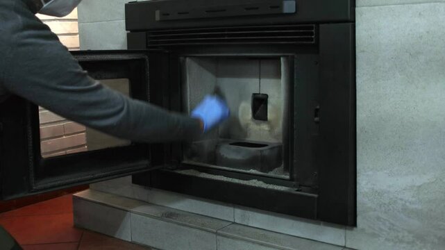 Man cleaning a pellet stove with a brush, wearing gloves and a protective mask. Indoor maintenance of a fireplace, ensuring hygiene and proper functioning in a cozy home setting.