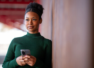 Thoughtful woman holding smartphone while leaning against wall