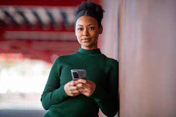 Woman holding smartphone while leaning against wall outdoors