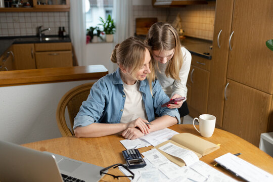 mother and daughter are sitting at the kitchen table in the kitchen. Papers, receipts, a laptop, a calculator are scattered on the table, creating the concept of family budgeting and joint financial p