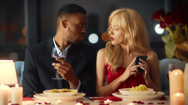In a cozy restaurant setting, a diverse couple enjoys their dinner. The young man attentively watches as the woman joyfully checks her phone, showcasing cheating in relationship