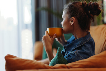 A woman sitting on a couch with a cup of coffee, looking out the window in contemplation