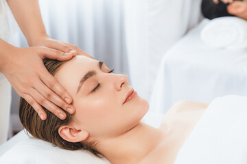 Caucasian couple enjoying relaxing anti-stress head massage and pampering facial beauty skin recreation leisure in dayspa modern light ambient at luxury resort or hotel spa salon. Quiescent