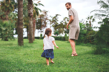 Fototapeta premium Father and his little daughter walking barefoot on the grass in a park, surrounded by trees, enjoying quality time and connecting with nature