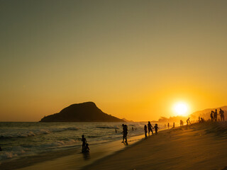 Pessoas aproveitando na baira do mar com p&ocirc;r do sol na praia com montanha ao fundo