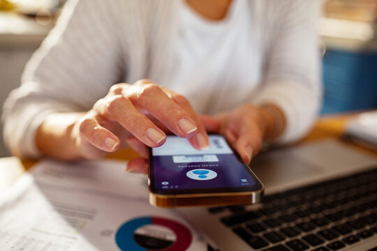 Senior woman using smartphone for online banking next to laptop at home