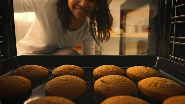 A millennial woman smiles as she peers into an oven filled with freshly baked cookies, enjoying a moment of home cooking in her bright kitchen. The warm atmosphere reflects her passion for baking.
