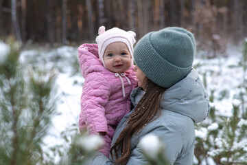 Mother holding her smiling baby daughter in a pink jacket in a snowy forest.