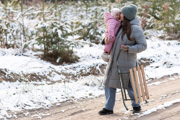 Mother walking in snowy forest with daughter in pink snowsuit and sled