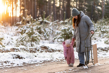 Mother walking in snowy forest with daughter in pink snowsuit and sled