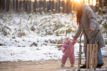 Mother walking in snowy forest with daughter in pink snowsuit and sled