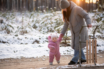 Mother walking in snowy forest with daughter in pink snowsuit and sled