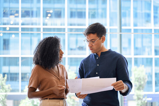Two young busy diverse coworkers, professional business man and business woman executive company team or employees working together standing in office discussing project documents. Candid photo.