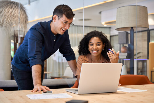 Two young busy diverse colleagues business people using laptop in modern office. Happy team employees working together on computer discussing online technology data solutions at work.