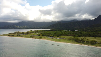 A drone captures the beauty of Kualoa Beach Park, showcasing golden sands, crystal-clear waves, and the dramatic Koʻolau Mountains towering above the serene Hawaiian coastline.