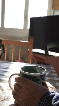 Old man's hands with smoky cup on dining table, indoor morning atmosphere at coffee time on winter day