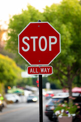 Red metal Stop All Way street sign on the side of a road, vertical, blurred green street background