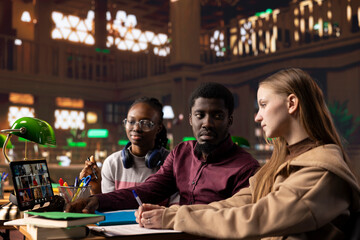 Young group of students engaging in a remote webinar course on tablet, participating at the online conference lesson from the university library. Diverse people learn from academic tutor.
