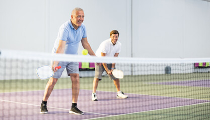 Focused resolved elderly man playing friendly pickleball match in team with male partner on indoors...