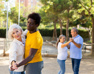 Fototapeta premium Exited older and middle-aged diverse people wearing casual clothes while enjoying latin dances breaking into pairs on sandy area between trees