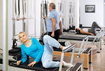 Fototapeta premium Focused elderly woman doing toning exercise for back on Pilates reformer bed in health fitness center