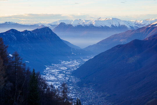 Snowy village is nestled in mountain valley at dusk, surrounded by towering peaks and mist, creating peaceful and remote alpine scene near Piani di Bobbio, Italy.