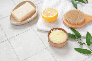 Bowl of lemon body scrub with massage brush, towel and soap on white tile background